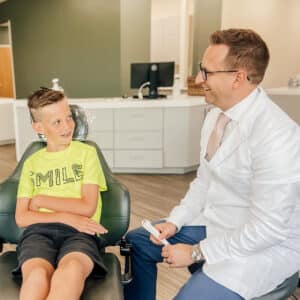 A child with braces sits in a dental chair, conversing happily with a person in a white coat, in a modern dental office.