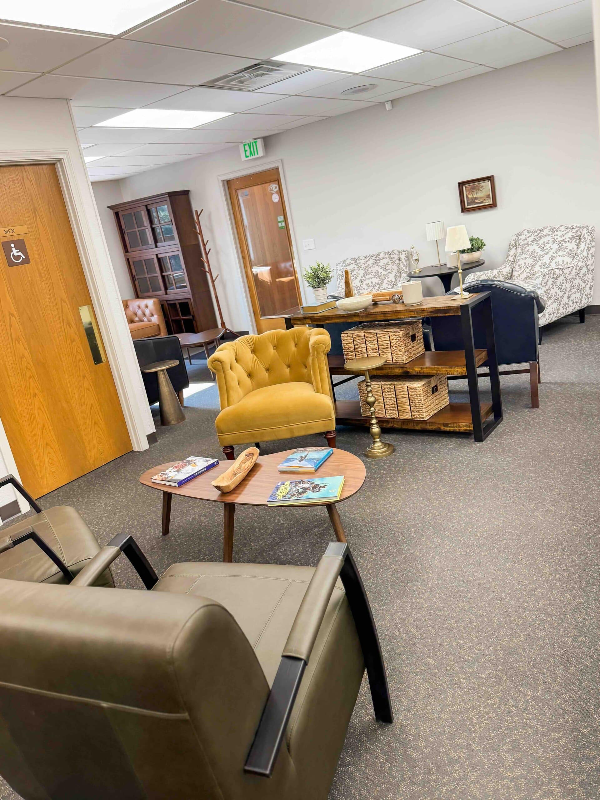 A cozy waiting room features armchairs, a small table with magazines, a tan armchair, bookshelves, decorative plants, and soft lighting for comfort.
