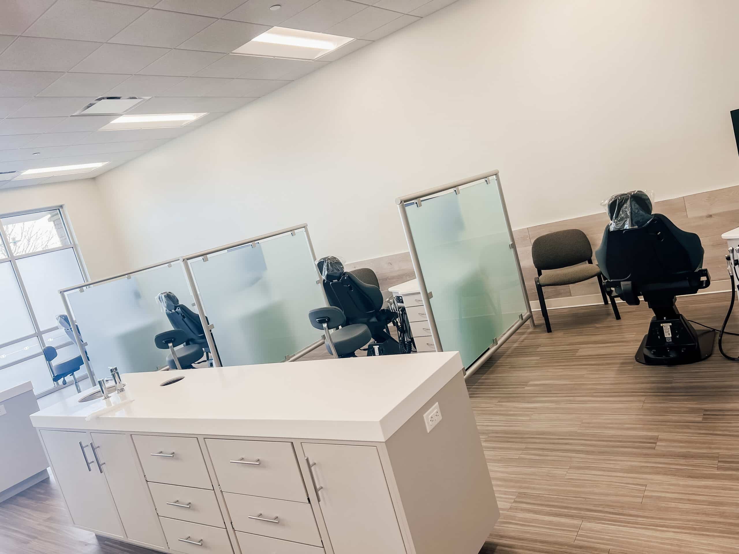Modern dental office with multiple dental chairs separated by frosted glass partitions, bright lighting, wooden flooring, and cabinets for storage.