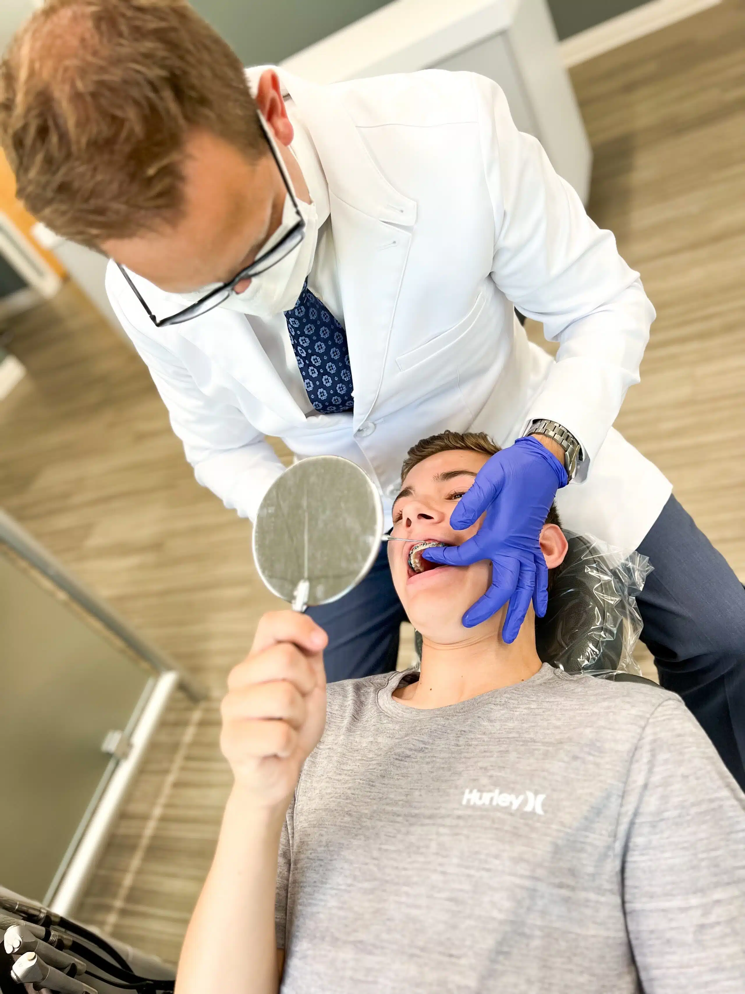 A person in a white coat examines another person's teeth with braces while they hold a mirror in a dental office.