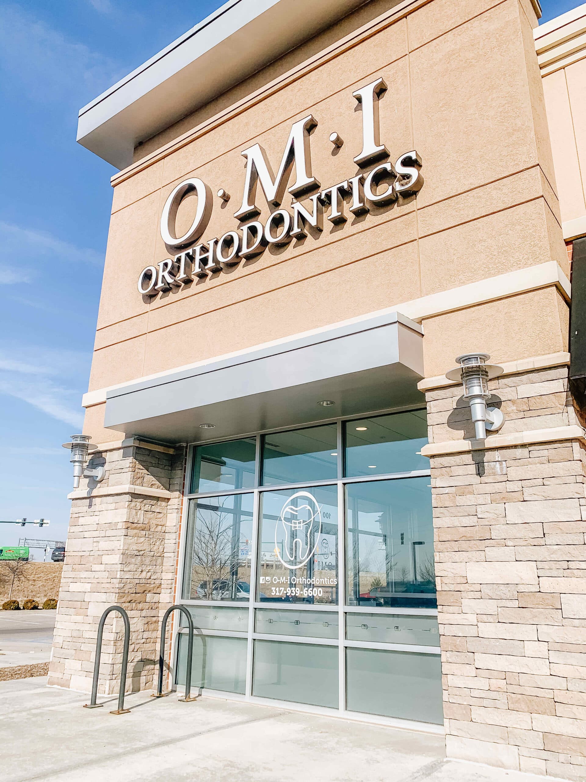 A dental clinic labeled "O.M.I Orthodontics" has a glass entrance, stone accents, and clear signage with contact information, under a clear blue sky.