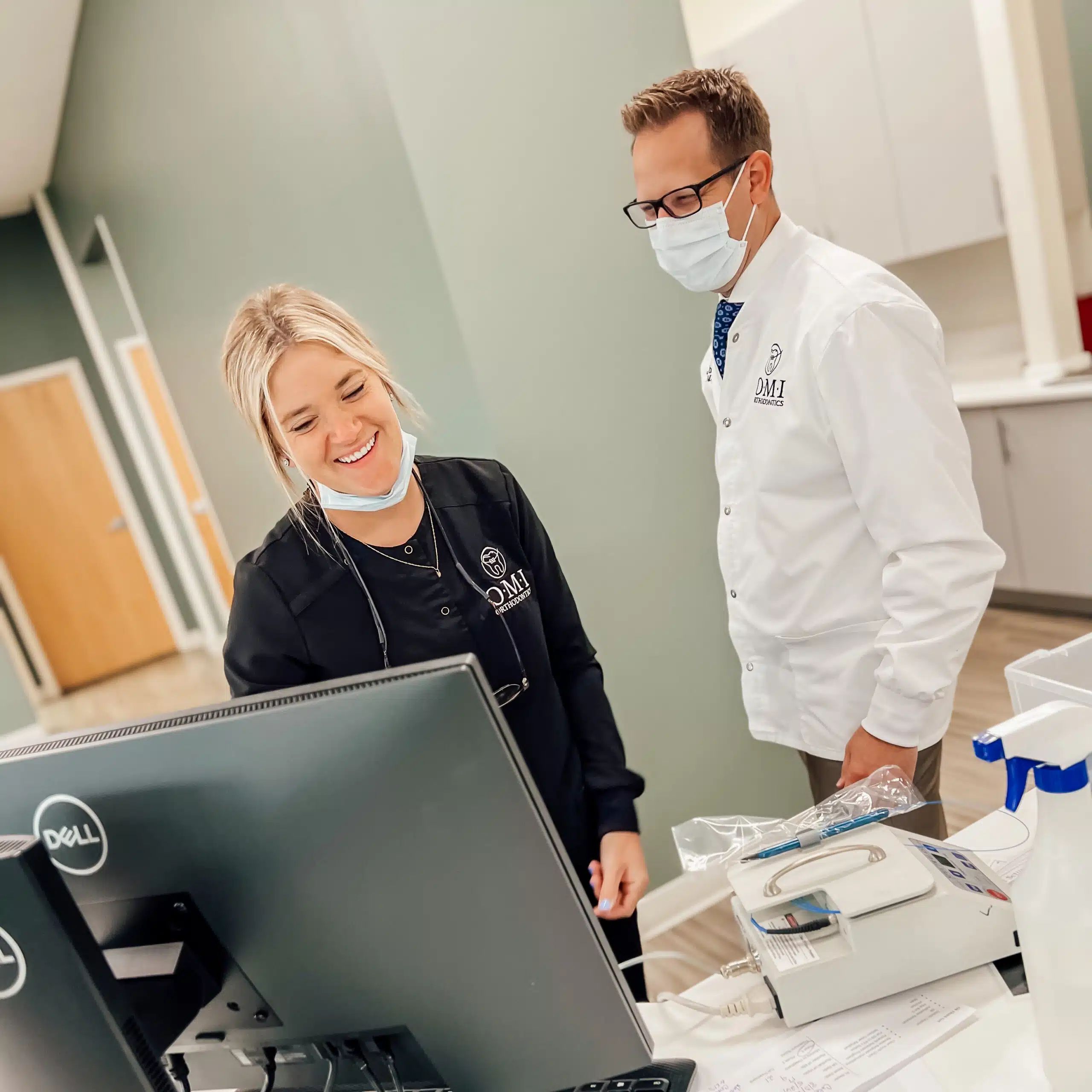 Two people with masks in a medical office, engage with a computer screen. One person wears a lab coat. No landmarks visible.