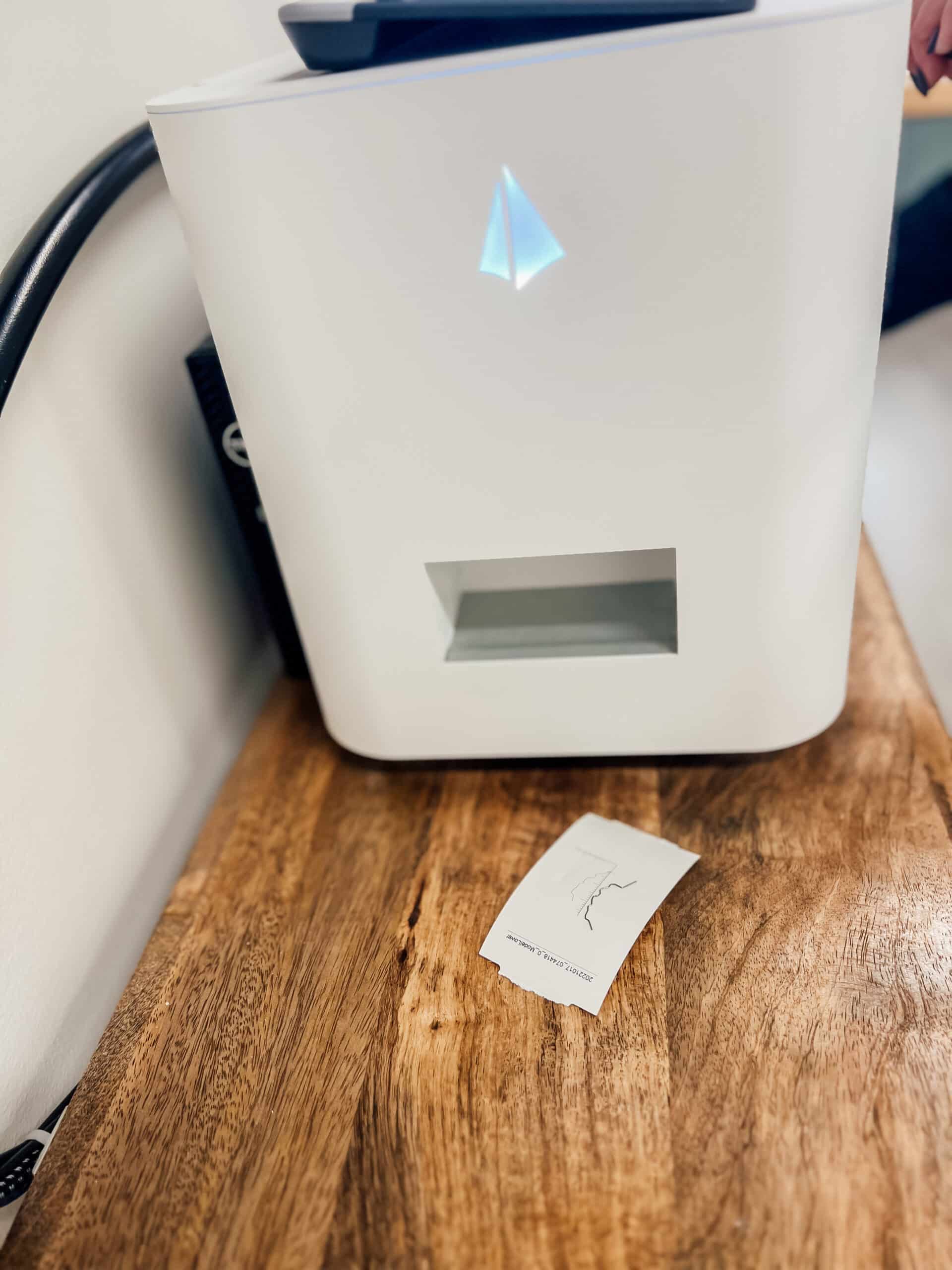 A white printer sits on a wooden desk, emitting a glowing blue light. A printed piece of paper lies nearby.