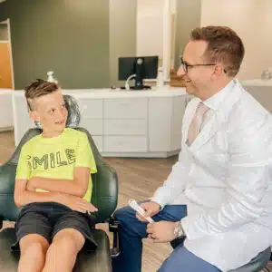 A child with braces sits in a dental chair, conversing happily with a person in a white coat, in a modern dental office.