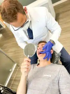 A person in a white coat examines another person's teeth with braces while they hold a mirror in a dental office.