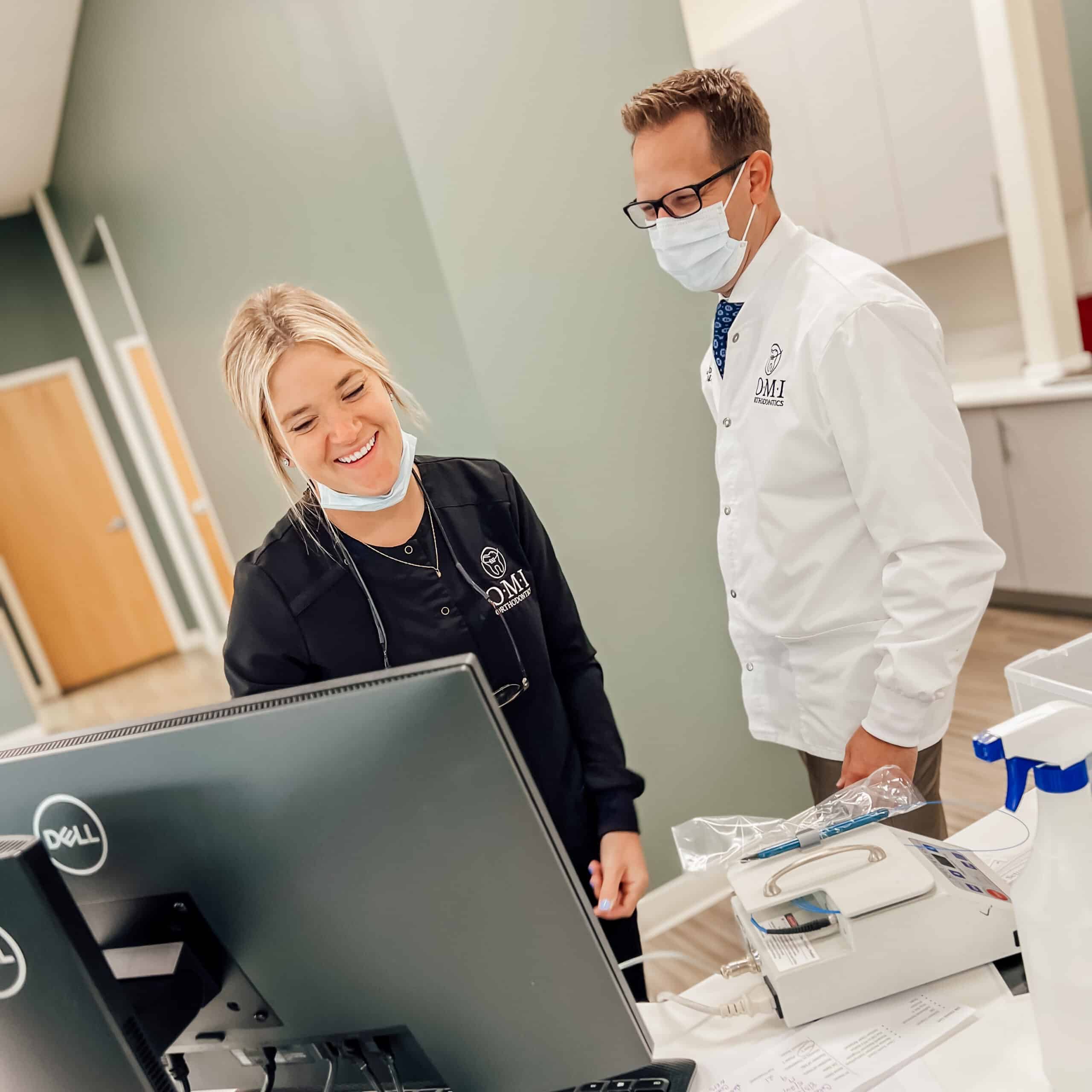 Two people with masks in a medical office, engage with a computer screen. One person wears a lab coat. No landmarks visible.