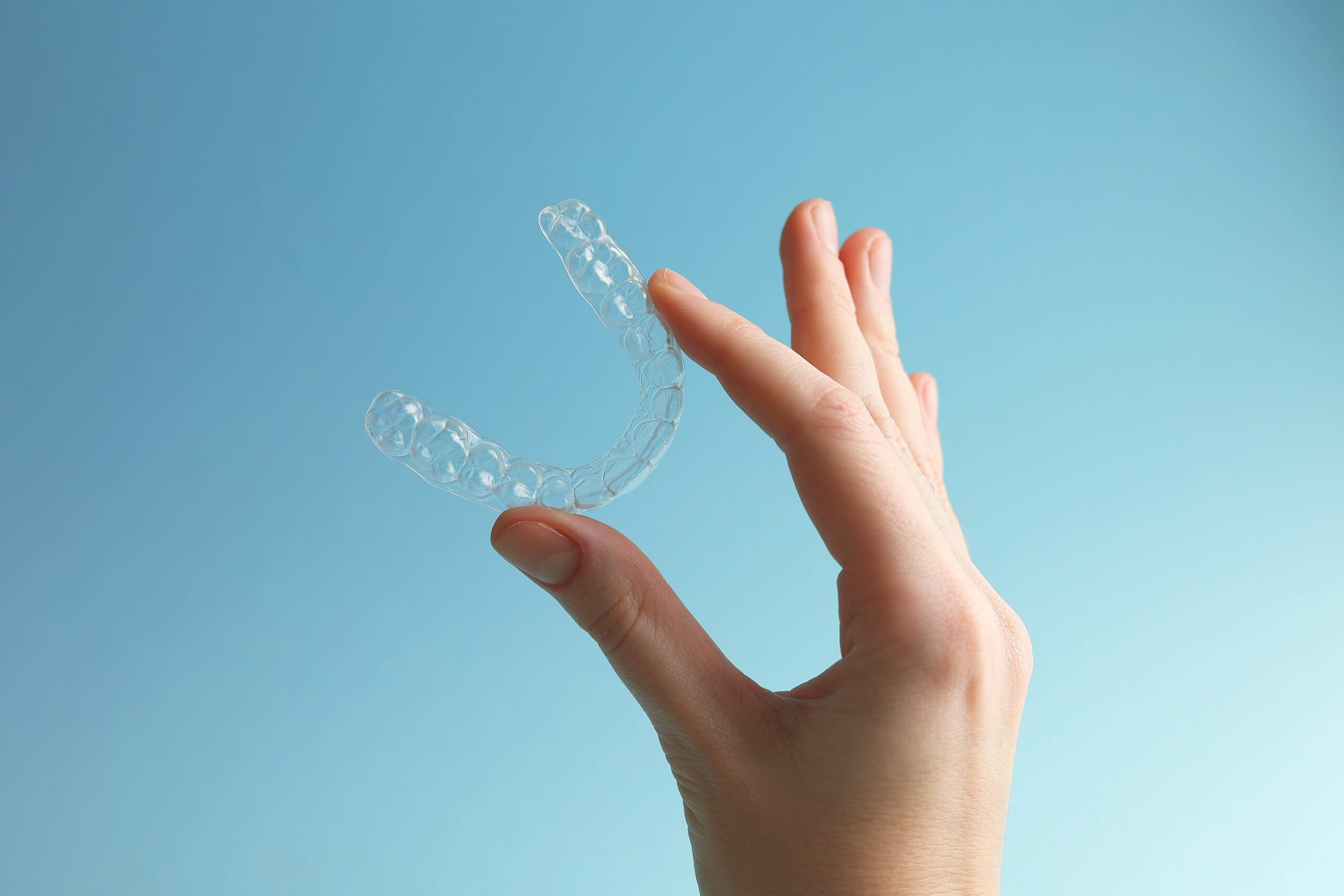A person’s hand holds a clear dental aligner against a blue background, demonstrating orthodontic equipment.