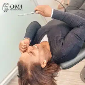 A person lies back in a dental chair, smiling with braces. Logo indicates it's an orthodontic setting.